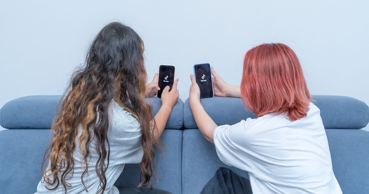 Duas jovens sentadas em um sofá, cada uma segurando um smartphone com o aplicativo TikTok aberto na tela. As duas estão posicionadas de costas para a câmera, uma com cabelos longos e ondulados, e a outra com cabelo curto tingido de vermelho.