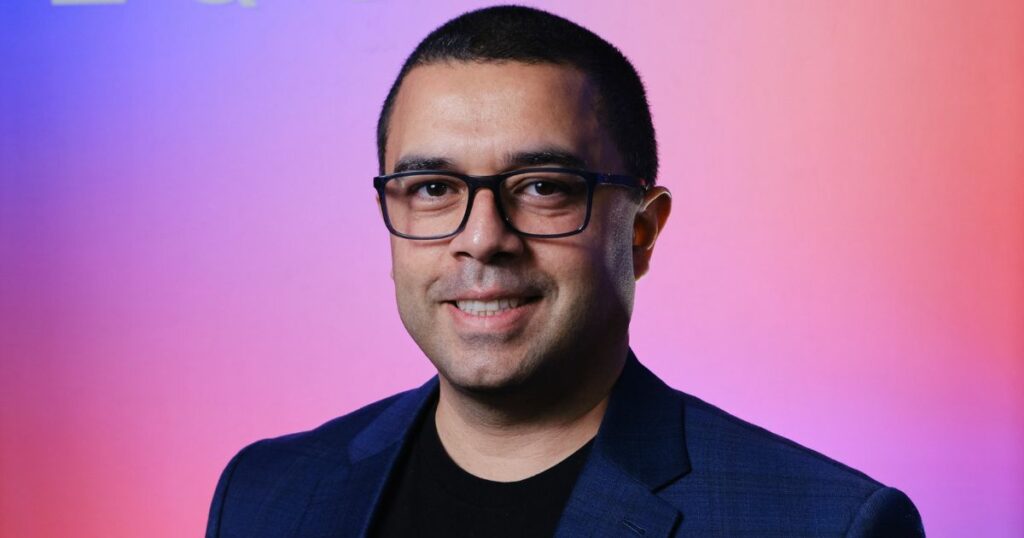 Victor Arnaud, executivo da Equinix, sorrindo para a câmera com um fundo de cores vibrantes em tons de roxo e rosa. Ele usa óculos e veste um blazer azul escuro sobre uma camiseta preta, transmitindo um visual moderno e profissional