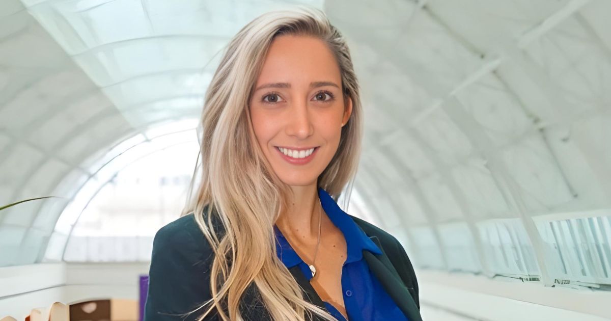 Foto de Andrea Quintana, executiva da Irani Ventures. Ela está sorrindo, com cabelos loiros e veste uma camisa azul sob um blazer escuro. O fundo exibe um ambiente moderno com arquitetura de linhas curvas e iluminação suave, destacando sua expressão acolhedora e profissional, startups