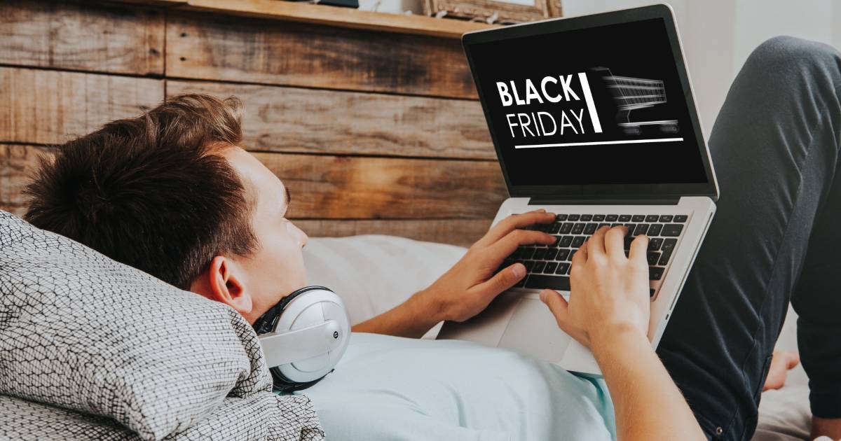 A imagem mostra uma pessoa relaxando em uma cama, usando um laptop e com fones de ouvido ao redor do pescoço. No laptop, é possível ver a tela com o texto "BLACK FRIDAY", indicando que a pessoa está navegando em ofertas da Black Friday. O ambiente ao fundo possui uma parede de madeira, criando uma atmosfera aconchegante. A pessoa está usando uma camiseta clara e parece focada na tela do computador enquanto aproveita as promoções