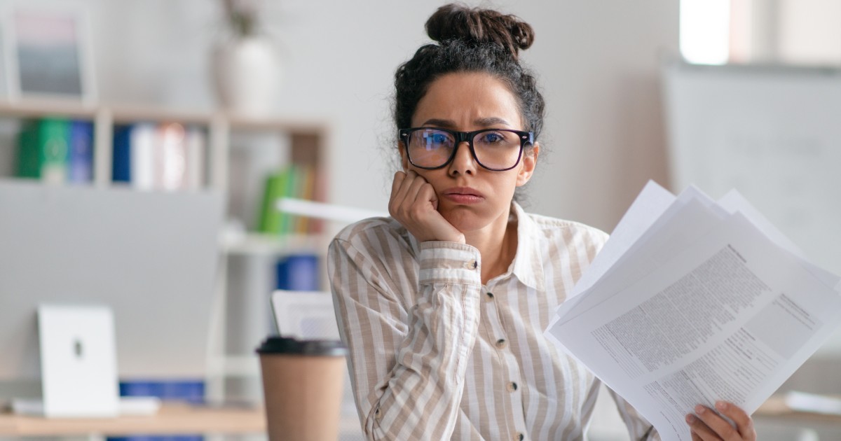Mulher com expressão de cansaço e preocupação, usando óculos e segurando papéis em um ambiente de escritório. Ela veste uma camisa listrada e está com a mão apoiada no rosto, transmitindo sensação de sobrecarga ou estresse no trabalho. Ao fundo, um espaço de trabalho desfocado com livros e objetos de escritório