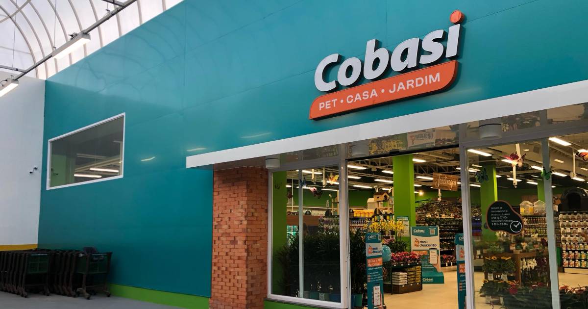 A imagem mostra a fachada de uma loja da Cobasi, uma rede de varejo especializada em produtos para pets, casa e jardim. A entrada é iluminada, com uma grande janela de vidro que revela o interior da loja, que parece ser organizada e bem decorada. O nome "Cobasi" está em destaque, em letras grandes e brancas, acompanhado dos termos "PET · CASA · JARDIM", com a palavra "Cobasi" destacada em laranja e verde. Em frente à loja, há alguns carrinhos de compras empilhados. A decoração interna parece ter várias prateleiras com produtos para pets e jardinagem