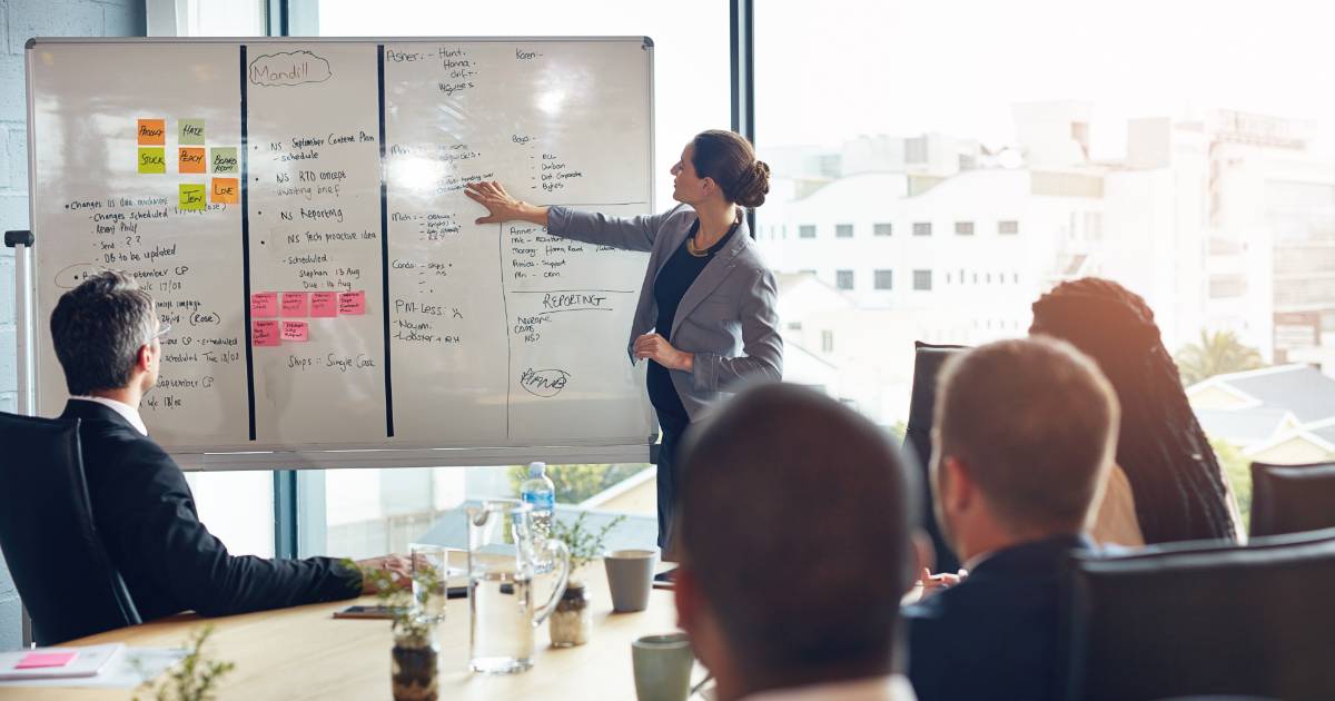 Imagem de uma reunião de negócios, onde uma mulher está em pé explicando estratégias em um quadro branco cheio de anotações e gráficos. Ao redor, um grupo de profissionais sentados escuta atentamente, sugerindo colaboração e planejamento estratégico em um ambiente corporativo, gestão de projetos