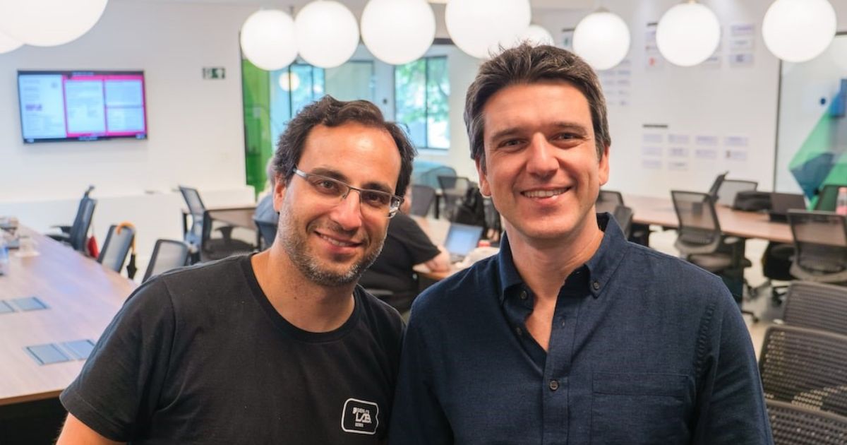 Gustavo Gierun e Gustavo Araújo, da Distrito, em um ambiente moderno de escritório, com mesas de trabalho ao fundo e uma atmosfera colaborativa iluminada por grandes luminárias brancas (gustavo gierun, gustavo araujo, distrito)