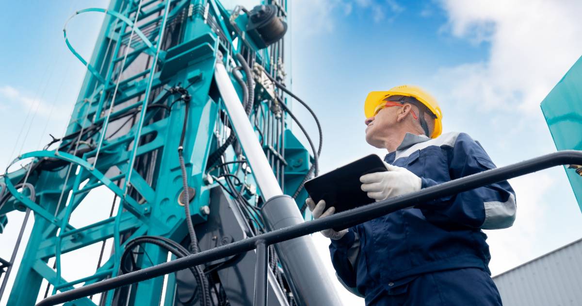 Trabalhador da indústria usando capacete amarelo e segurando um tablet enquanto inspeciona uma grande máquina industrial azul. Ele veste uniforme de proteção e luvas brancas. O céu azul ao fundo complementa o ambiente industrial ao ar livre, política, indústria