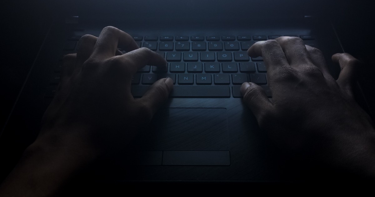 Mãos digitando em um teclado de laptop em um ambiente escuro, iluminadas por uma luz fraca que destaca o cenário misterioso e sombrio. A imagem evoca o conceito do lado obscuro da tecnologia, como cibercrimes ou atividades clandestinas, valor