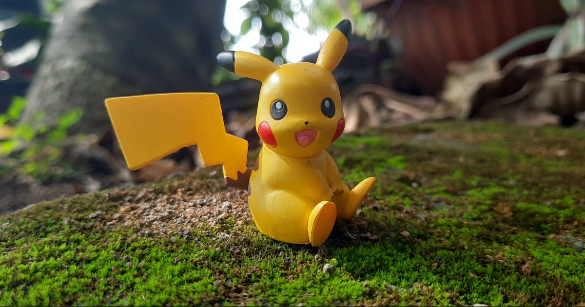 Figura de um Pikachu, pokémon, em miniatura, sentado sobre musgo em um ambiente natural, com um fundo desfocado de plantas e árvores.