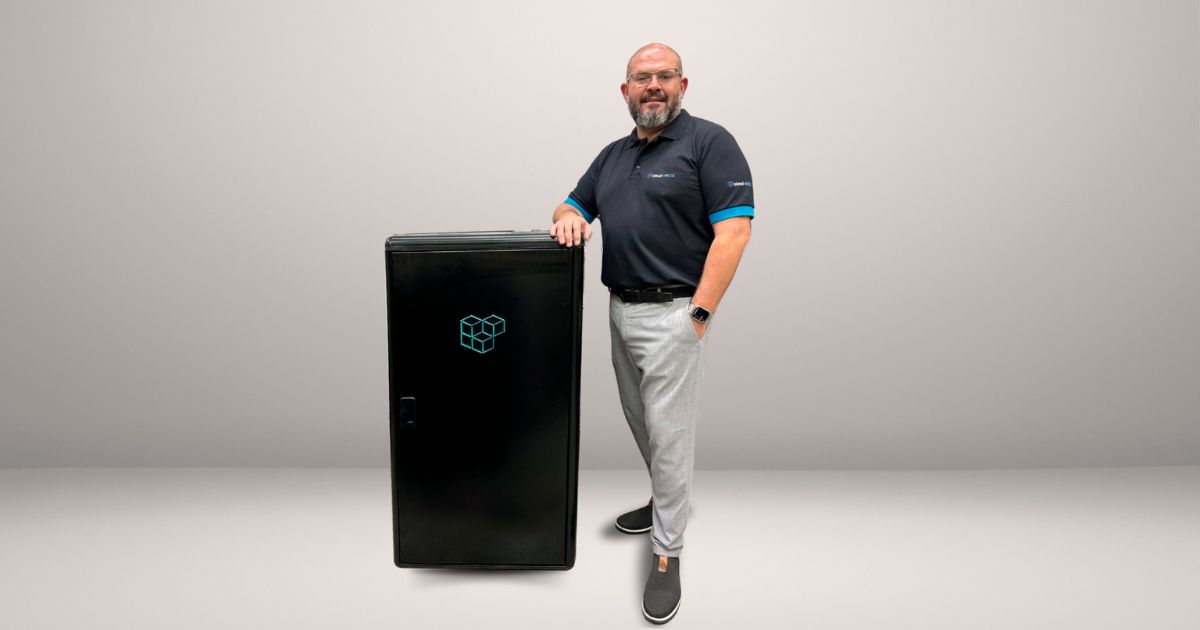 A imagem apresenta André Machado, head de pesquisa, desenvolvimento e inovação da cloudvBOX, posando ao lado do vBOX EDGE, a solução desenvolvida pela empresa. Ele está vestido com uma camisa polo preta, com detalhes em azul nas mangas e o logotipo da empresa, além de calça clara e sapatos casuais escuros. O vBOX EDGE, exibido ao seu lado, é um equipamento de cor preta com o logotipo da cloudvBOX em destaque, ilustrando o design compacto e funcional do micro data center. O fundo é neutro, direcionando toda a atenção para André e o equipamento.