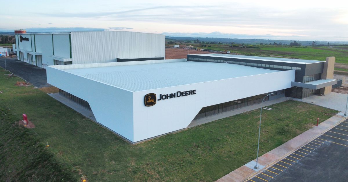 Vista aérea de uma unidade da John Deere localizada em uma área ampla, com design moderno em tons de branco e o logotipo da empresa na fachada. O local está cercado por áreas verdes e estacionamentos, com um horizonte que revela paisagens rurais ao fundo (CPD-I, john deere, indaiatuba, P&D)