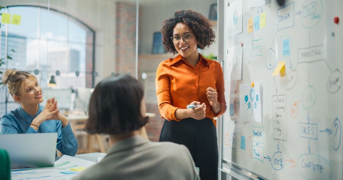 A imagem mostra um ambiente de escritório moderno, onde três pessoas estão em uma reunião colaborativa. No centro, uma mulher está de pé, vestindo uma camisa laranja e óculos, explicando ou apresentando algo em um quadro branco cheio de diagramas, post-its e anotações. Ela parece estar liderando a discussão com entusiasmo. À esquerda, uma mulher loira, vestindo azul, está sentada e sorrindo, demonstrando atenção e engajamento. À direita, outra pessoa está parcialmente visível de costas, também participando da reunião. O ambiente é iluminado, com janelas grandes ao fundo, criando uma atmosfera acolhedora e profissional.