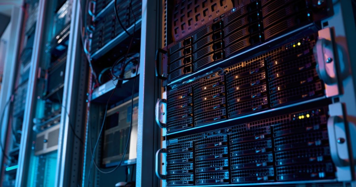 A imagem mostra um ambiente de data center com diversos servidores montados em racks. Os equipamentos estão organizados em fileiras verticais, com luzes indicadoras acesas em alguns dos dispositivos, sugerindo que estão em operação. A iluminação é predominantemente azulada, com tons sutis de roxo, criando uma atmosfera tecnológica e moderna. Cabos e componentes conectam os servidores, refletindo a infraestrutura robusta necessária para armazenamento e processamento de dados em larga escala. Esse cenário é típico de locais utilizados para operações de TI críticas, como hospedagem de sites, análise de dados, inteligência artificial e outras aplicações de computação avançada. (emissões, brasil)