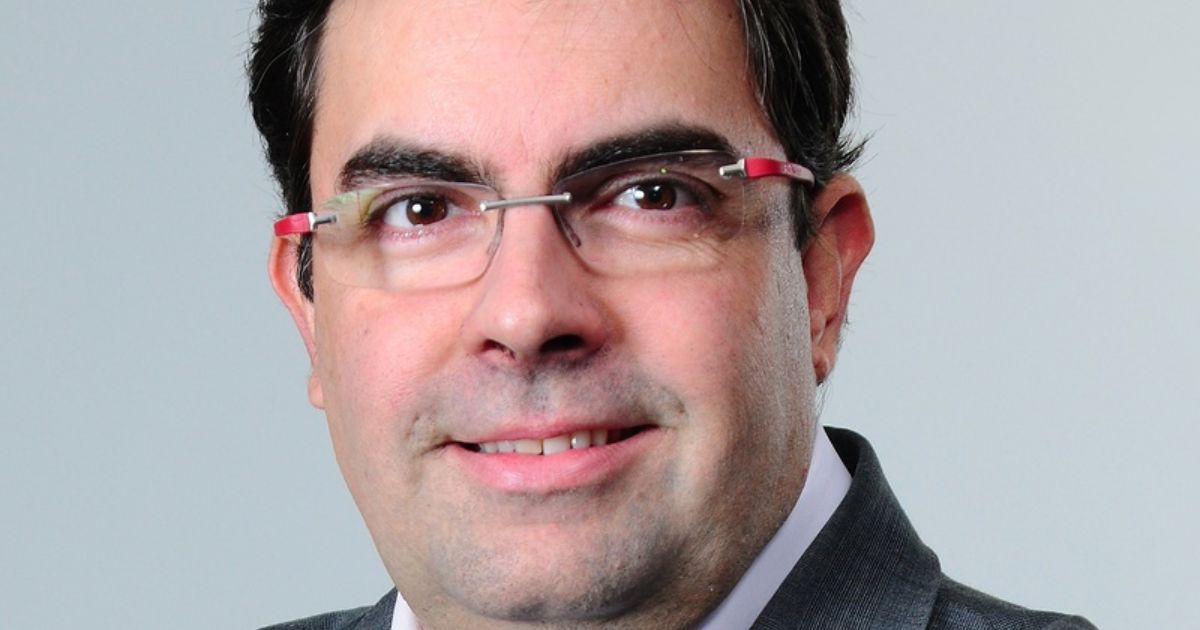 A imagem mostra um homem com cabelo castanho escuro, curto, e usando óculos com armação de borda fina e detalhes vermelhos. Ele está vestido formalmente, usando um blazer cinzento sobre uma camisa branca. A expressão facial é amigável, com um leve sorriso. O fundo da imagem é neutro, de cor cinza clara, destacando a pessoa no retrato (Alberto Claro, EY)