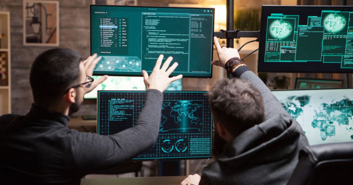 A imagem mostra dois profissionais de tecnologia observando atentamente múltiplas telas de computador. Cada monitor exibe gráficos, códigos e informações relacionadas à cibersegurança, com destaque para mapas globais e dados em tempo real. Eles parecem estar analisando informações de forma colaborativa, apontando para detalhes específicos nos monitores. A atmosfera da cena é focada e intensa, com os profissionais imersos em seu trabalho, refletindo um ambiente de monitoramento ou investigação digital.