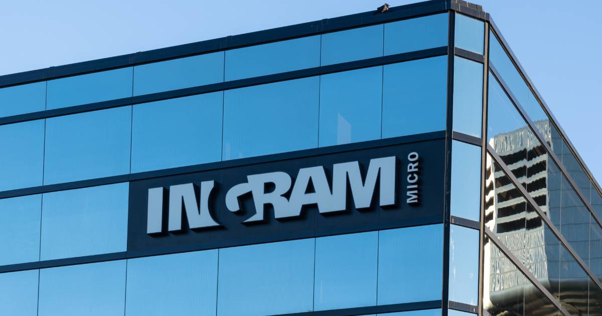 Logotipo da Ingram Micro em destaque na fachada de um prédio corporativo com janelas de vidro reflexivas, que refletem o céu azul e partes de outros edifícios ao fundo. A logo está localizada no canto superior de um edifício moderno