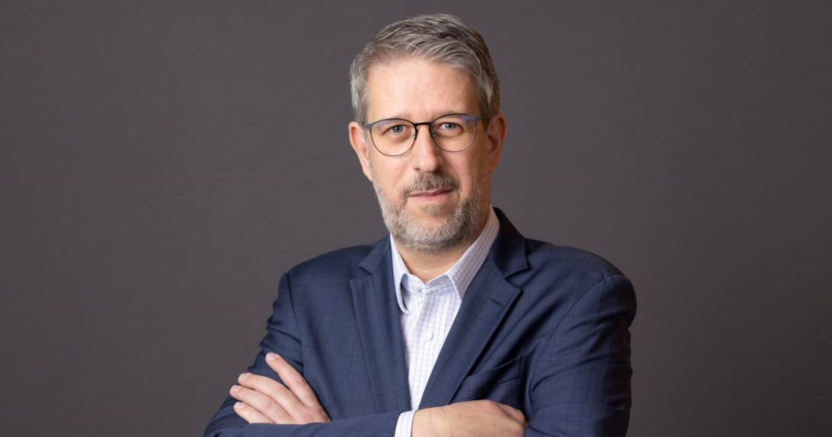 Na foto, Juliano de Conti aparece posicionado diante de um fundo neutro, vestindo um paletó azul-escuro sobre uma camisa social clara. Ele mantém os braços cruzados e exibe uma expressão séria e confiante, típica de um executivo seguro de suas decisões. Seu olhar direcionado à câmera transmite profissionalismo e compostura, reforçando a imagem de liderança e competência.