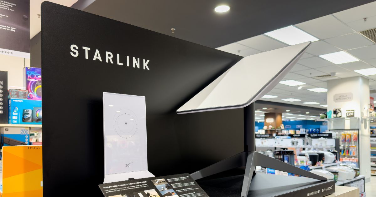 Uma exibição de um produto Starlink em uma loja de eletrônicos. No centro da imagem, há um painel preto com o logotipo 'Starlink' em letras brancas. Um terminal de usuário Starlink está em destaque, com a antena parabólica moderna e angular posicionada sobre um suporte robusto. À esquerda, está a unidade do roteador branco com o logotipo da SpaceX. Ao fundo, é possível ver prateleiras e outros produtos eletrônicos disponíveis na loja, sob luzes de teto brilhantes. (Anatel)