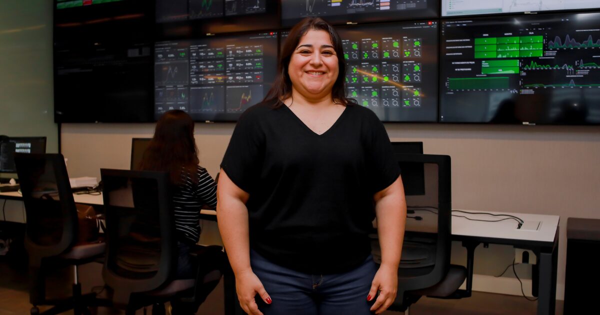 Vanessa Bustamante, CIO da HDI Seguros, sorri enquanto está em pé no centro de operações da empresa, com painéis digitais de monitoramento ao fundo.
