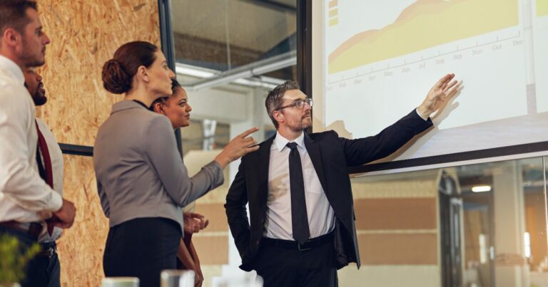 Executivo em uma sala de reuniões apontando para um gráfico projetado em uma tela, enquanto apresenta dados de controladoria para um grupo de pessoas. O ambiente é moderno, com detalhes de madeira e vidro, indicando um espaço corporativo. (produto, liderança, líder, julgamentos)