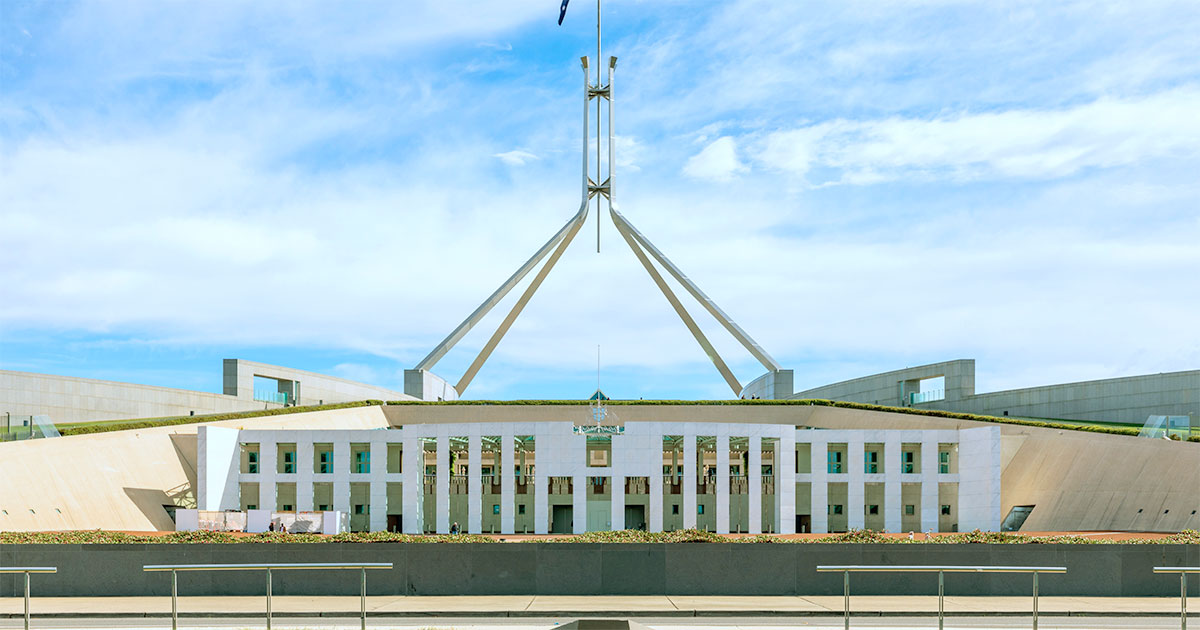 Fachada do Parlamento Australiano em Canberra, destacando sua arquitetura moderna com linhas simétricas, colunas brancas e um grande mastro central com a bandeira da Austrália. O céu azul com nuvens complementa a vista frontal do edifício, redes sociais