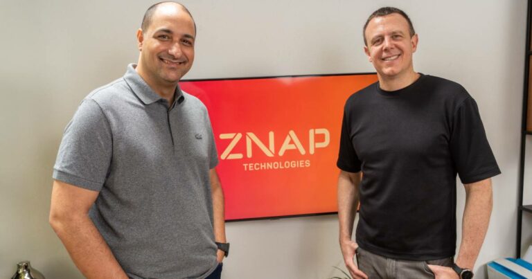 Luciano de Medeiros, CTO, e Ricardo Junqueira, CEO, da ZNAP Technologies, posam sorrindo em frente a um monitor que exibe o logotipo da empresa em fundo vermelho. Ambos estão em um ambiente moderno, transmitindo profissionalismo e inovação
