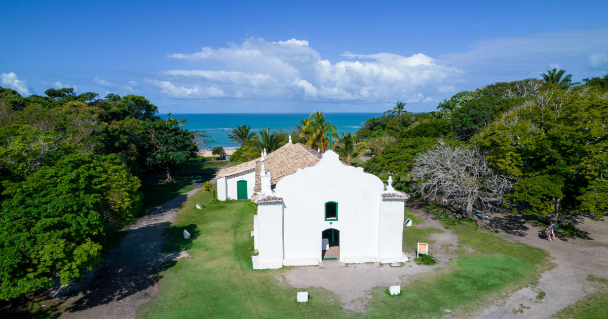Vista aérea da Igreja de São João Batista, localizada em Trancoso, com o mar ao fundo e rodeada por árvores. O céu está claro, com algumas nuvens, e o ambiente transmite tranquilidade e beleza natural, IT Forum Trancoso