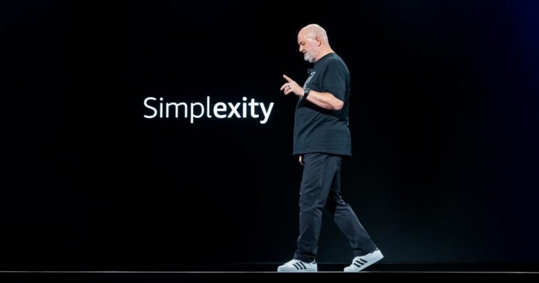 Werner Vogels, CTO da Amazon, em uma apresentação no palco durante o re:Invent, o principal evento da Amazon Web Services (AWS). Ele caminha enquanto gesticula, ao lado da palavra "Simplexity" projetada em um fundo preto, representando o equilíbrio entre simplicidade e complexidade na inovação tecnológica. Com seu estilo casual, usando uma camiseta preta e tênis Adidas, Vogels reflete sua abordagem direta e prática ao discutir o futuro da tecnologia e seu impacto no mundo.