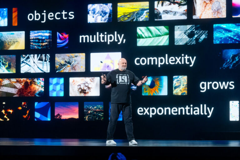 A imagem mostra Werner Vogels, CTO da Amazon, em um grande palco no re:Invent 2024, em Las Vegas, vestido de forma casual com camiseta preta e calça escura, apresentando em frente a um painel digital. O painel exibe várias imagens coloridas e texturas diversificadas, acompanhadas por palavras como "objects", "multiply", "complexity", "grows", "exponentially". A disposição visual sugere um tema relacionado à tecnologia, crescimento exponencial de dados ou complexidade no ambiente digital. O palestrante gesticula enquanto explica, conferindo dinamismo à apresentação. O cenário é iluminado por tons vibrantes e o fundo escuro destaca o conteúdo projetado e o apresentador.
