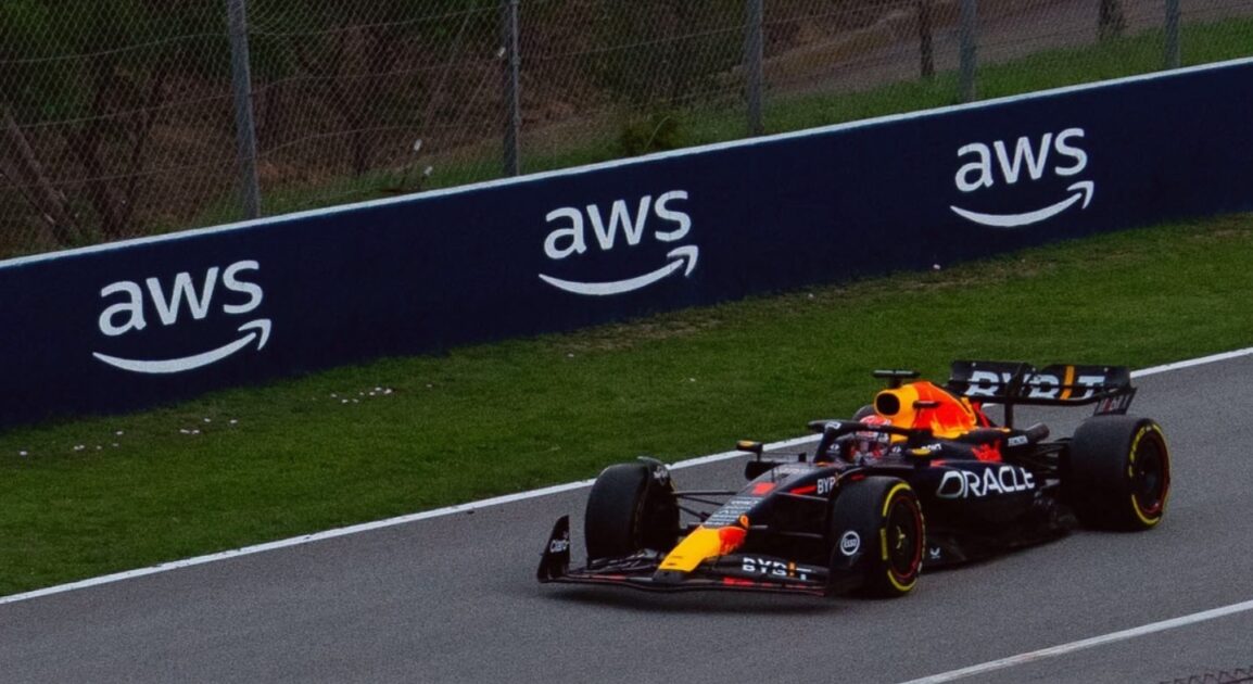 Carro de Fórmula 1 da equipe Oracle Red Bull Racing em alta velocidade em uma pista de corrida, com placas de publicidade da AWS visíveis ao longo do circuito. A imagem destaca o dinamismo e a tecnologia no automobilismo