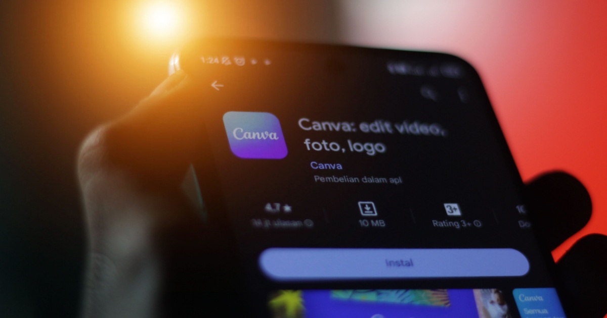 Close-up de uma mão segurando um smartphone exibindo a página de instalação do aplicativo Canva, com informações como tamanho do app, classificação e opção para instalar. Fundo com iluminação em tons de laranja e vermelho
