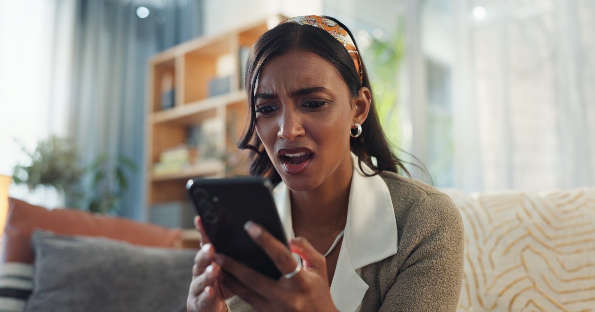 Mulher expressando surpresa ou preocupação enquanto olha para o celular, retratando o impacto de possíveis notícias ou informações inesperadas no ambiente doméstico, desinformação, fake news