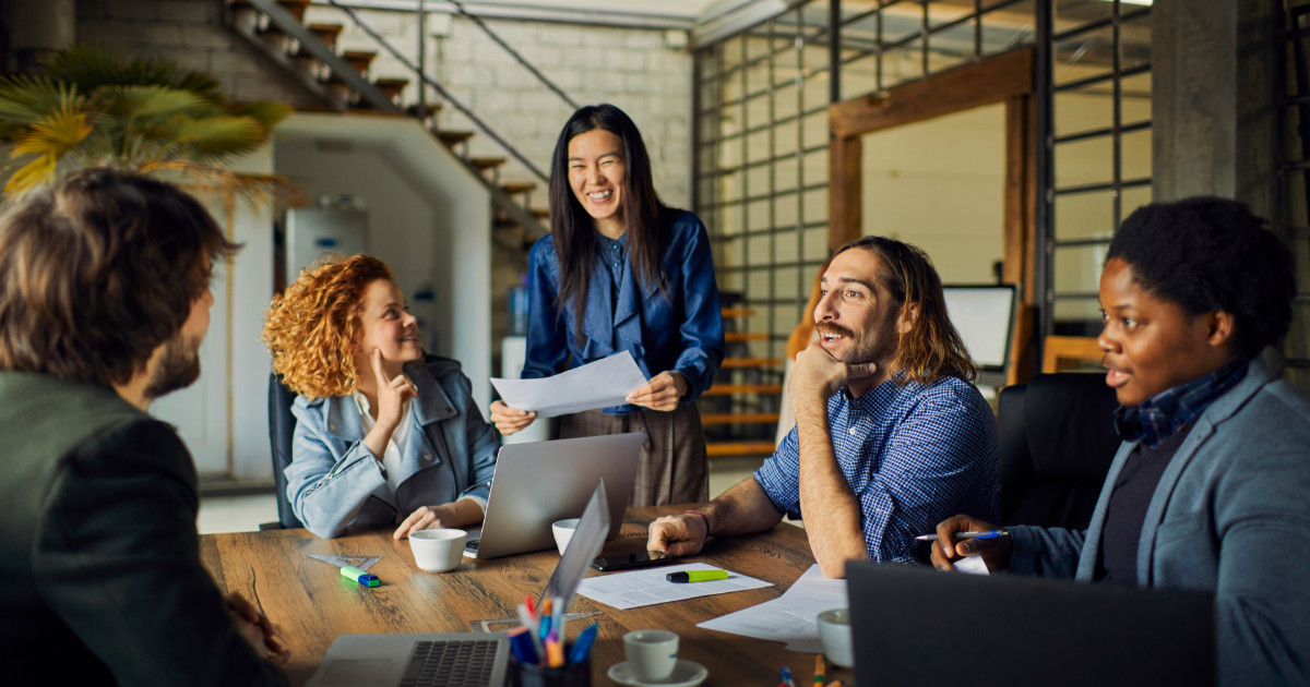 A imagem mostra um grupo diverso de profissionais em uma reunião colaborativa. Cinco pessoas estão sentadas ao redor de uma mesa, sorrindo e interagindo. Uma mulher está em pé, segurando alguns papéis, enquanto os outros estão ouvindo atentamente e sorrindo. Há duas mulheres e três homens, representando uma diversidade de etnias e estilos. A mesa está organizada com laptops, canetas, e papéis, criando um ambiente descontraído e produtivo. O ambiente é moderno, com iluminação natural e uma parede de tijolos ao fundo, sugerindo um espaço criativo de trabalho (diversidade, ento)