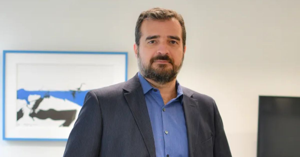Foto de Fabio Câmara, fundador e CEO do Grupo FCamara, vestindo um terno azul escuro e camisa azul clara. Ele está em um ambiente corporativo com uma obra de arte emoldurada ao fundo, transmitindo um ar profissional e confiante