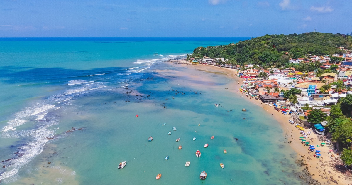 Vista aérea da Praia de Pipa, no Rio Grande do Norte, com mar azul claro, pequenas embarcações na água e a faixa de areia ocupada por turistas e guarda-sóis coloridos. Ao fundo, uma vila com casas coloridas e áreas verdes