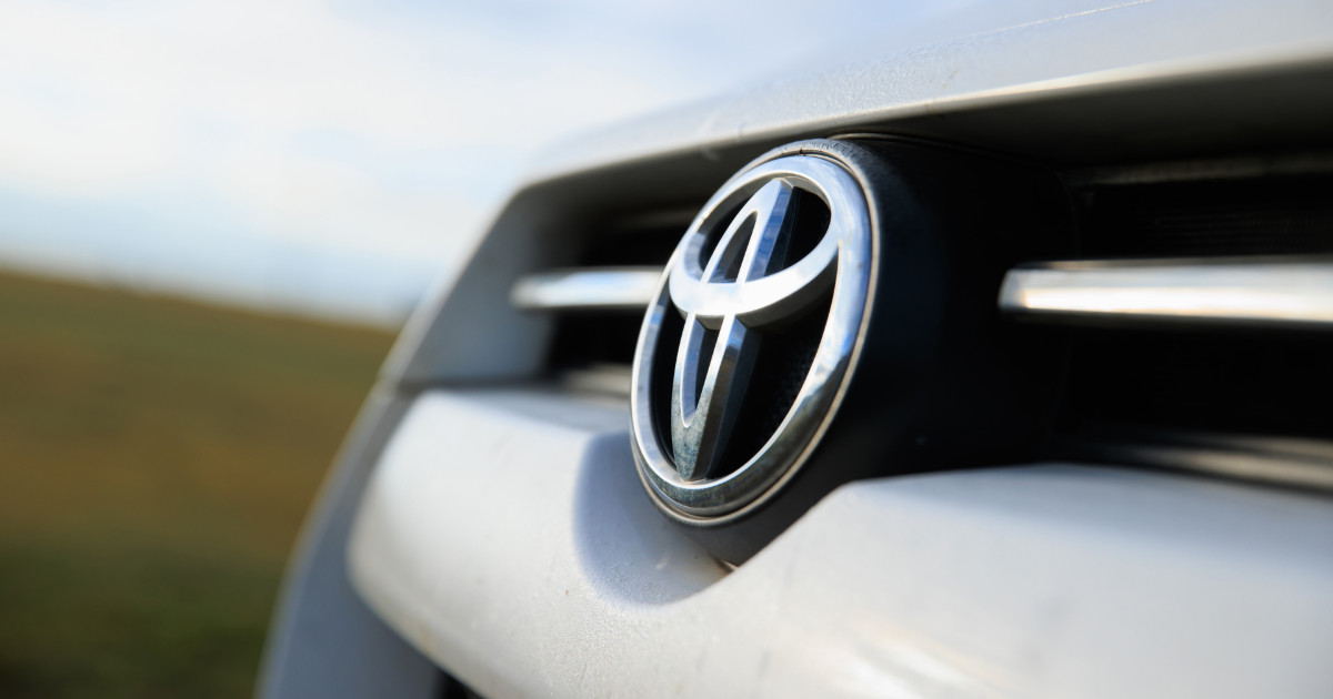 Close-up do emblema da Toyota na grade frontal de um veículo, destacando o logotipo prateado em alto relevo sobre fundo escuro. Ao fundo, desfocado, uma paisagem natural com céu claro