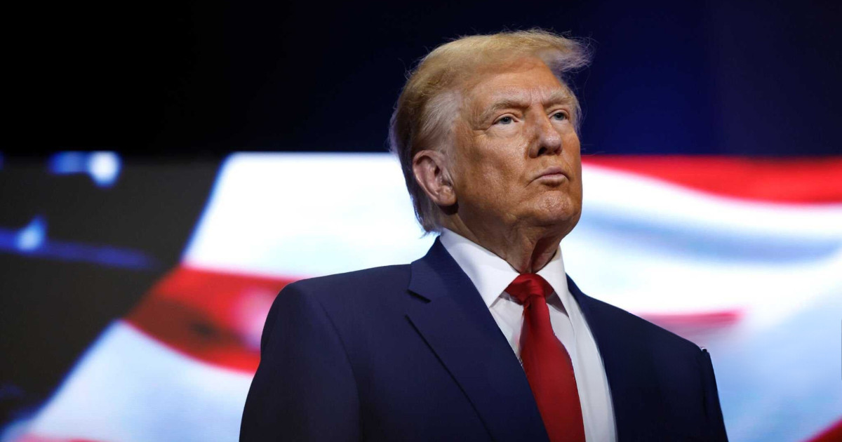 Donald Trump, presidente dos Estados Unidos, em destaque. Ele está usando um terno azul escuro, camisa branca e gravata vermelha, com uma expressão séria e olhar fixo. Ao fundo, há um padrão desfocado em EUA vermelho, branco e azul, remetendo às cores da bandeira americana. A imagem transmite formalidade e autoridade (ordem executiva, ações)