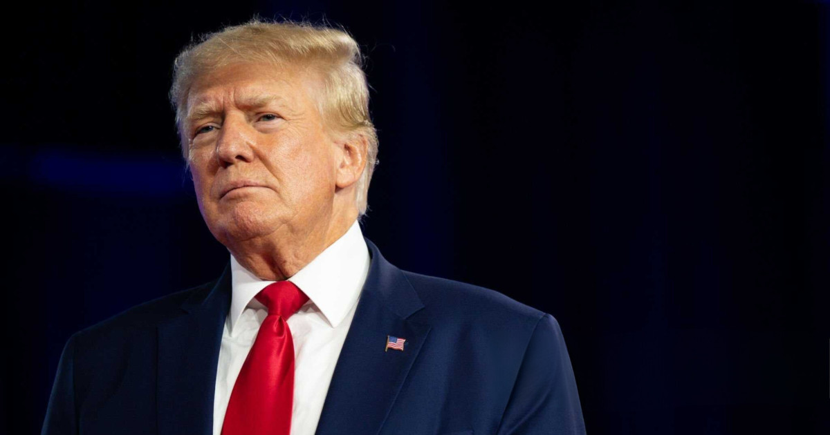 Donald Trump, presidente dos Estados Unidos, em destaque. Ele está usando um terno azul escuro, camisa branca e gravata vermelha, com um pin da bandeira americana na lapela. Sua expressão é séria, e o fundo escuro realça sua figura. eletronica