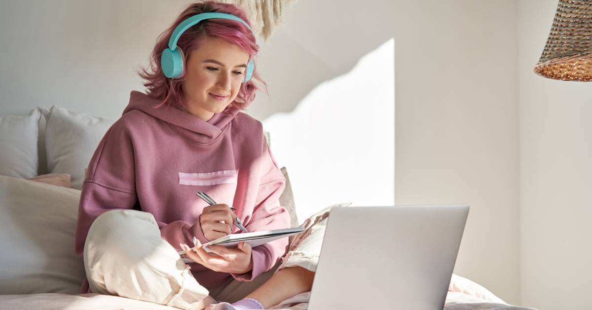 Jovem com cabelo rosa usando fones de ouvido azuis, sentada em um ambiente aconchegante, enquanto escreve em um caderno e olha para a tela de um laptop. A cena transmite um momento de estudo ou trabalho remoto com tecnologia
