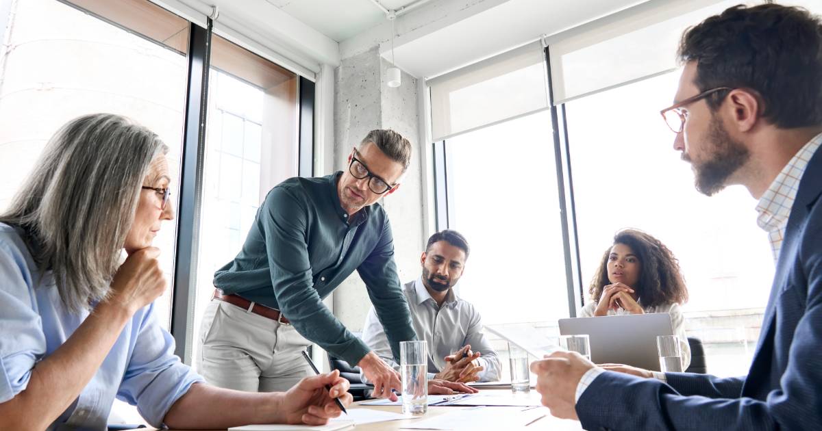 Grupo de profissionais em uma reunião de trabalho em um ambiente corporativo moderno, com janelas amplas ao fundo. Um homem está em pé, gesticulando e apontando para documentos na mesa, enquanto os demais participantes escutam atentamente, discutindo estratégias ou tomada de decisões (gestão de projetos)