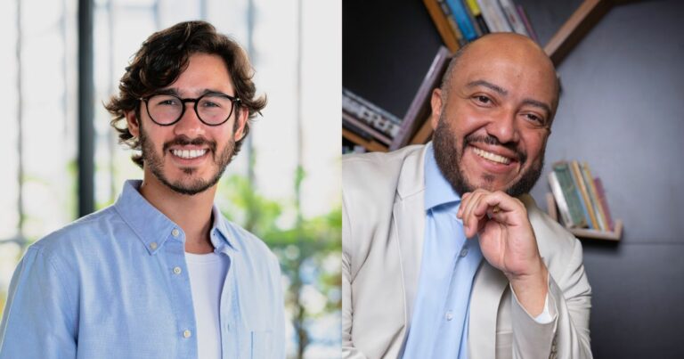 Duas pessoas, ambas usando óculos, estão lado a lado. À esquerda, um homem jovem com barba curta, cabelo ondulado e uma camisa azul clara. À direita, um homem mais velho com terno e gravata azul, cabelo curto e uma expressão formal. O fundo é dividido: claro à esquerda, com janelas, e escuro à direita, com luzes de evento.