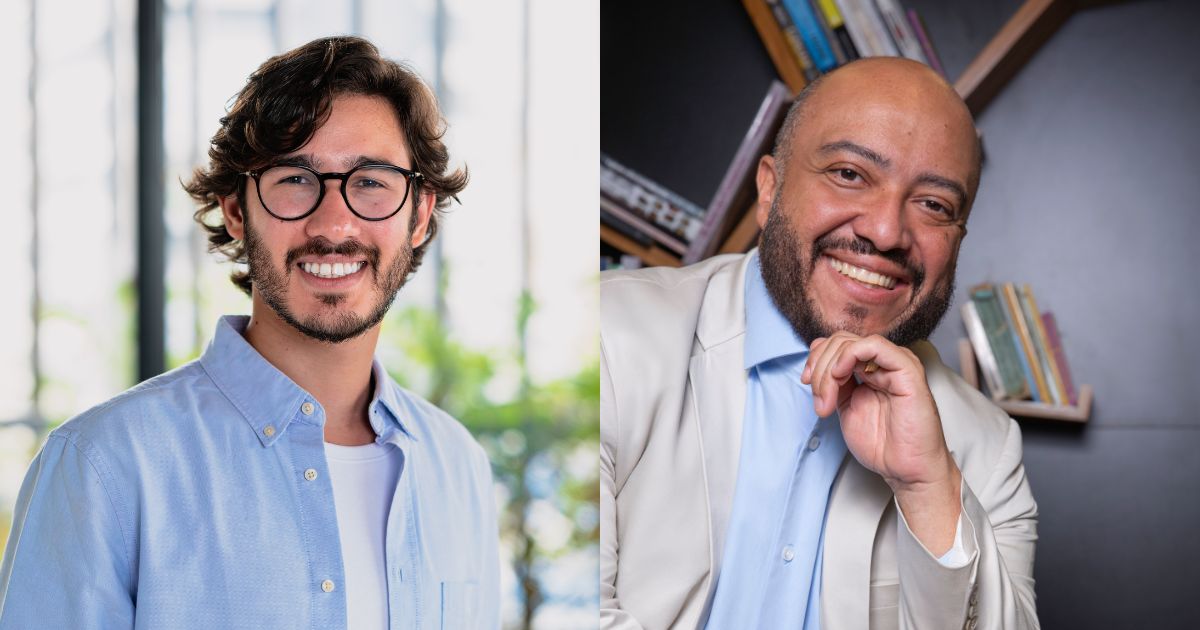 Duas pessoas, ambas usando óculos, estão lado a lado. À esquerda, um homem jovem com barba curta, cabelo ondulado e uma camisa azul clara. À direita, um homem mais velho com terno e gravata azul, cabelo curto e uma expressão formal. O fundo é dividido: claro à esquerda, com janelas, e escuro à direita, com luzes de evento.