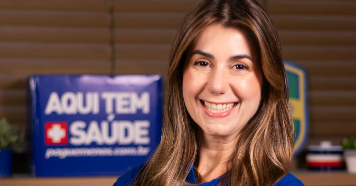 Priscila Braga, nova diretora de Digital e Inteligência de Vendas da Pague Menos, sorrindo em um ambiente corporativo. Ao fundo, um cartaz azul com os dizeres 'Aqui tem saúde' reforça a identidade da empresa