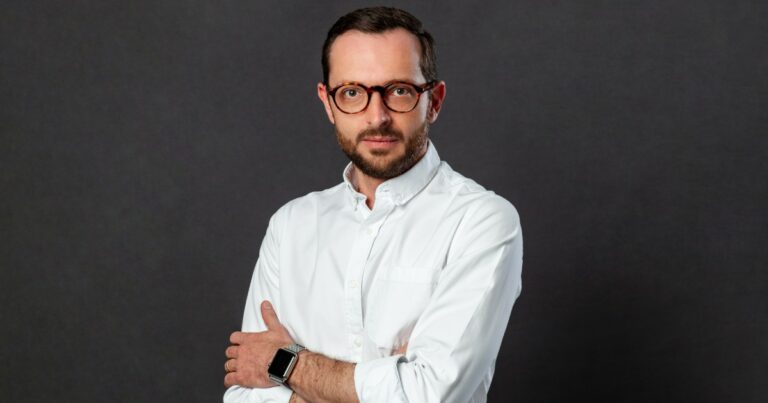 Rodrigo Tozzi, head de Operações da Tools for Humanity no Brasil, em um retrato formal com fundo cinza. Ele veste uma camisa branca, usa óculos de aro redondo e exibe uma expressão séria e profissional.