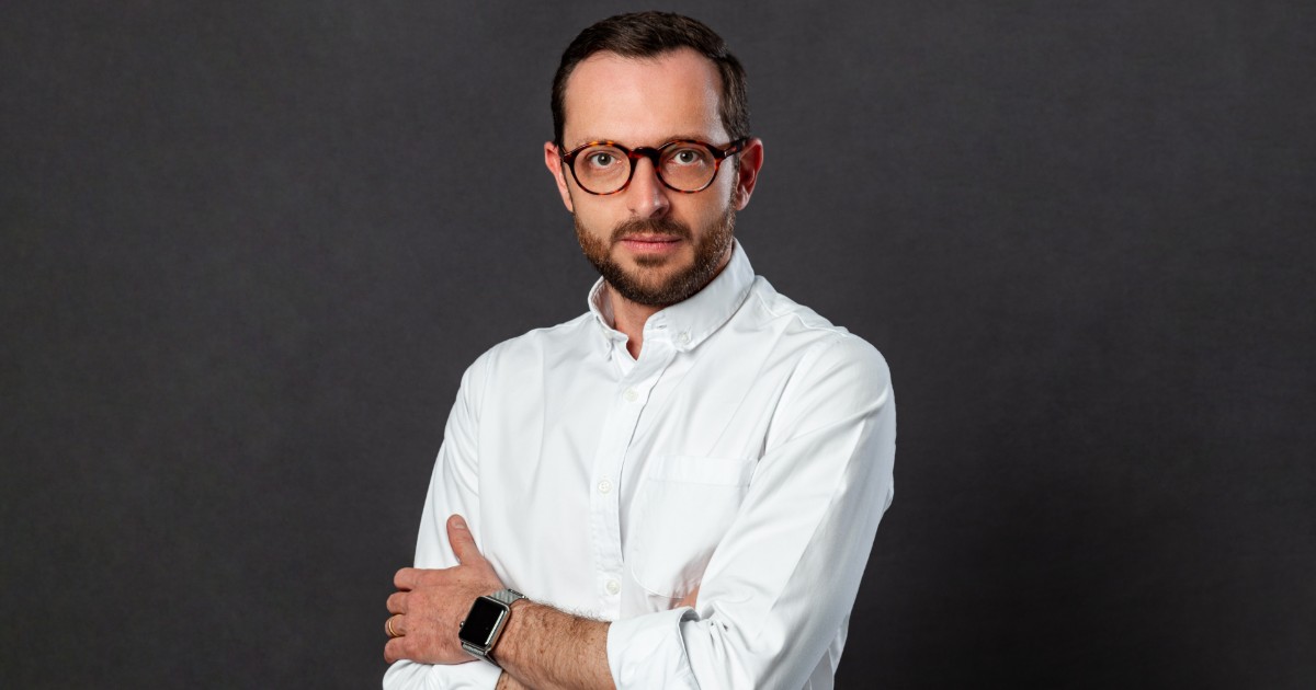 Rodrigo Tozzi, head de Operações da Tools for Humanity no Brasil, em um retrato formal com fundo cinza. Ele veste uma camisa branca, usa óculos de aro redondo e exibe uma expressão séria e profissional.