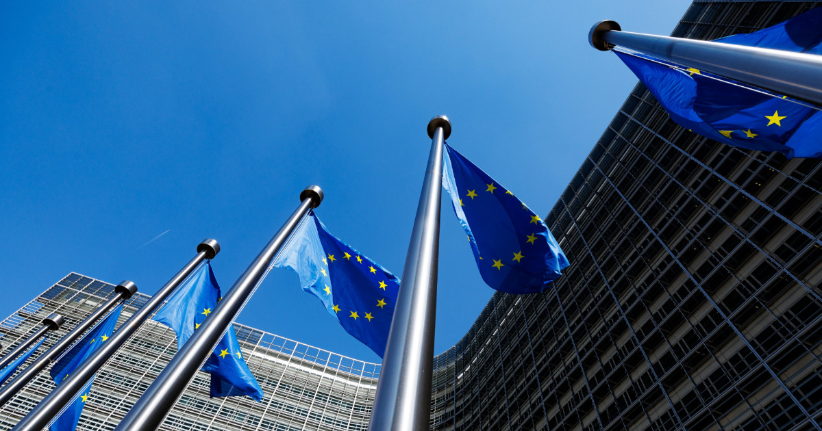 Bandeiras da União Europeia hasteadas em mastros prateados, tremulando contra um céu azul claro. Ao fundo, há um edifício moderno com fachada de vidro, representando instituições europeias. A imagem simboliza a união e cooperação entre os países-membros do bloco europeu. geopolitica