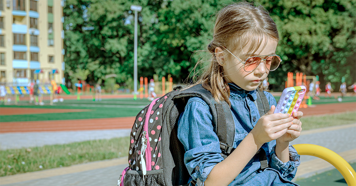 Uma criança sentada em um parque escolar usando óculos de sol e vestindo uma mochila de bolinhas nas costas. Ela está concentrada em um celular com uma capinha colorida. Ao fundo, há um campo de grama sintética com equipamentos de playground e árvores, compondo um ambiente de lazer ao ar livre (escolas)