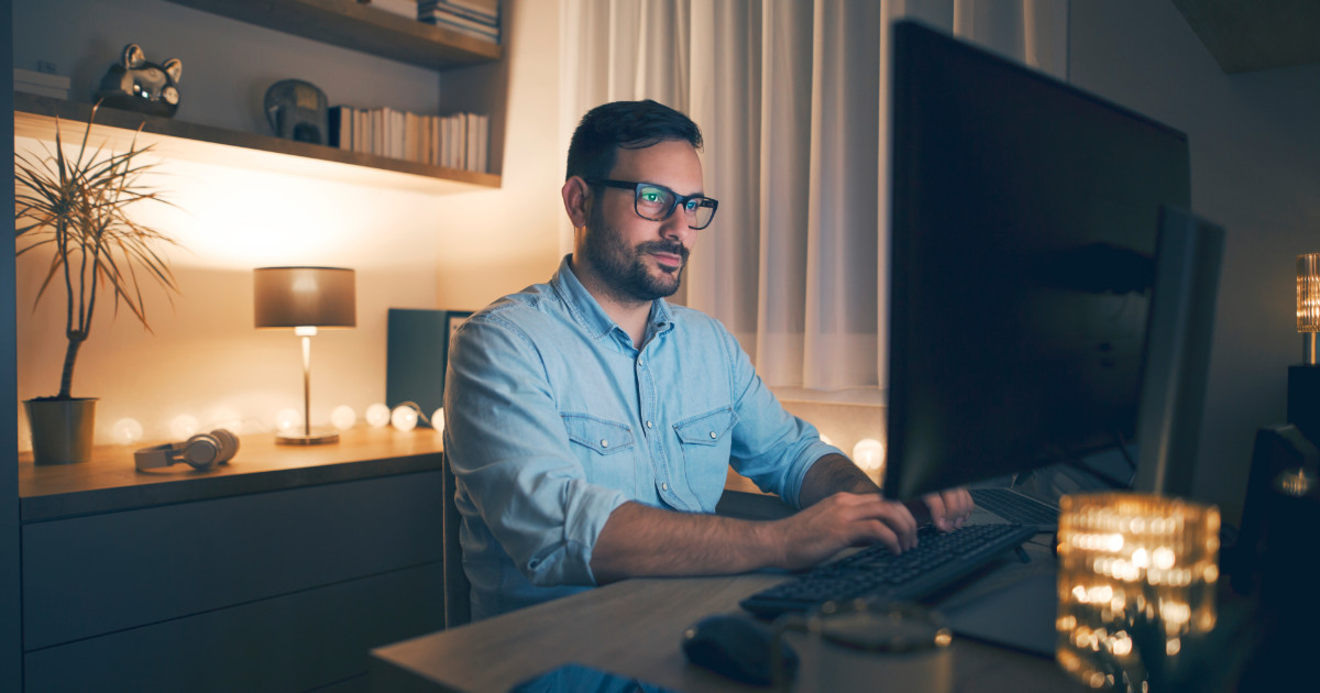 Homem usando óculos, sentado em frente a um computador, trabalhando em um ambiente aconchegante e bem iluminado. O local tem uma decoração moderna, com luminárias, plantas, estante de livros e itens decorativos ao fundo. A luz suave cria uma atmosfera confortável, ideal para trabalho remoto ou estudo (Robert Half, trabalho remoto, presencial, home office)