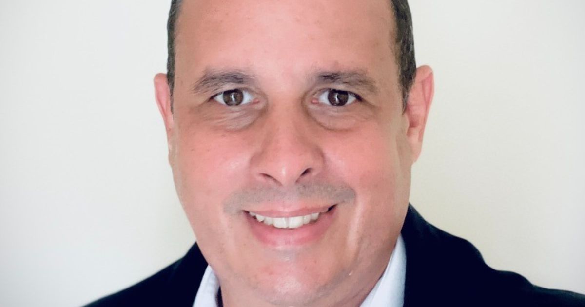 Imagem de Flavio Campos, novo head de TI da Coca-Cola Andina Brasil. Ele aparece sorrindo, com pele clara e cabelo curto escuro, vestindo um paletó preto sobre uma camisa branca. O fundo é neutro e claro, destacando seu rosto. A foto tem um tom profissional, adequada para apresentação corporativa