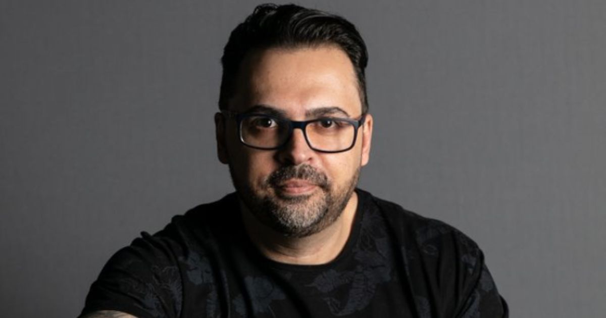 Texto alternativo: Retrato de Alexandre Bastos Borges, novo CIO da Pernambucanas. Ele é um homem de pele clara, barba curta e cabelo escuro penteado para trás. Usa óculos de armação preta e uma camisa preta com estampa discreta. O fundo é cinza neutro, e a iluminação destaca seu rosto.