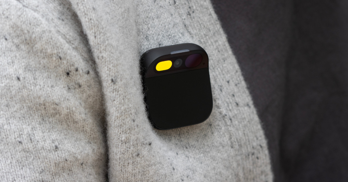 A imagem mostra um Humane Ai Pin, um dispositivo tecnológico compacto e vestível, fixado na roupa de uma pessoa. O aparelho tem um design quadrado com cantos arredondados e uma superfície preta fosca. Na parte superior, há uma seção que abriga sensores, câmeras e uma luz amarela acesa, indicando que o dispositivo está ativo. Ele está preso a um tecido de lã clara com pequenas manchas escuras, destacando seu design minimalista e discreto.