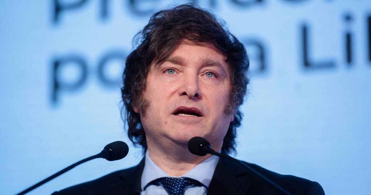 Javier Milei, presidente da Argentina, discursando em um evento. Ele aparece entre dois microfones, vestindo um terno escuro, camisa clara e gravata azul. O fundo exibe um telão com palavras parcialmente visíveis (cripto)